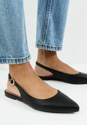 Pieds chaussés de ballerines slingback noires en cuir pointues avec des boucles dorées, associées à un jean denim bleu clair.