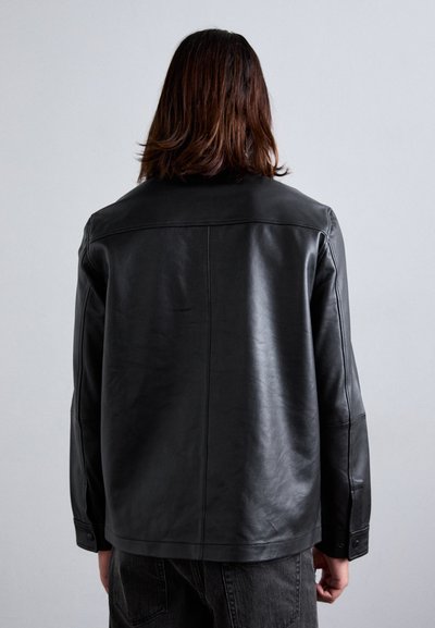 Person with shoulder-length hair wearing a black leather jacket and dark jeans, standing against a plain light gray background.