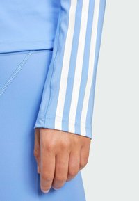Ljusblå atletisk tröja med vita tre-stripes detaljer på ärmen. Slät tyg med en åtsittande design och synliga sömnadsdetaljer.