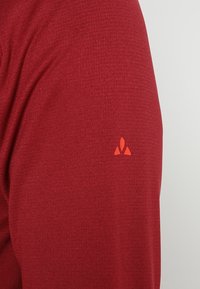 Tela de camiseta deportiva roja con una textura suave. Presenta un pequeño logo naranja en la manga izquierda, diseño minimalista.
