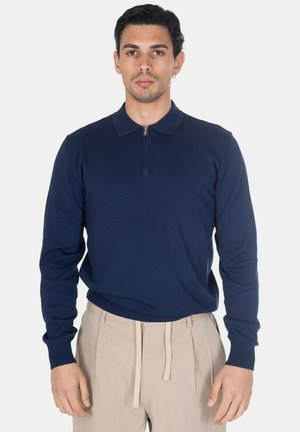 Uomo che indossa una polo blu navy a maniche lunghe con cerniera e pantaloni beige con coulisse, in piedi davanti a uno sfondo bianco.