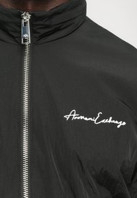 Sort letvægtsjakke med højt krave, sølvfarvet lynlås lukning og broderet "Armani Exchange" logo i hvidt på venstre side.