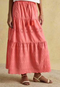 Fustă maxi în culoarea coral vibrant, cu detalii din ochiuri și tiv ondulat, având talie cu șnur și mici ciucuri. Asortată cu sandale joase.