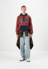 Un jeune homme portant un sweat à capuche maroon et navy de GAP, un jean baggy bleu clair, des baskets blanches et une veste marron nouée autour de sa taille.