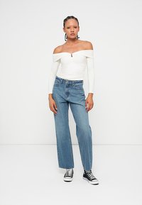 Off-shoulder white top with button detail, paired with high-waisted blue jeans and black sneakers. Casual, relaxed fit and design.