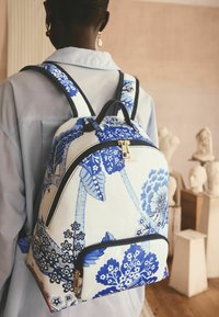 Mochila hecha de tela crema con patrones florales azules, que presenta herrajes negros, un bolsillo frontal y correas ajustables.