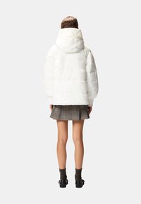 Giacca imbottita bianca con cappuccio, design trapuntato e maniche larghe. Indossata con una gonna a quadri e scarpe nere, mostrando un look a strati.