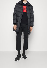 Svart puffjacka med dragkedja och hög krage, bärs över en svart sweatshirt med en röd grafik. Matchas med svarta cropped byxor och sneakers.