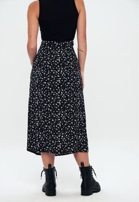 Mujer vestida con una blusa negra sin mangas, una falda midi negra con flores, botas de tobillo negras con cordones y un brazalete de oro, de pie contra un fondo blanco.