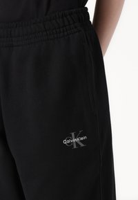 Čierne bavlnené tepláky s elastickým pásom. Na ľavej strane sa nachádza malé strieborné logo Calvin Klein. Hladká textúra, ležérny dizajn.