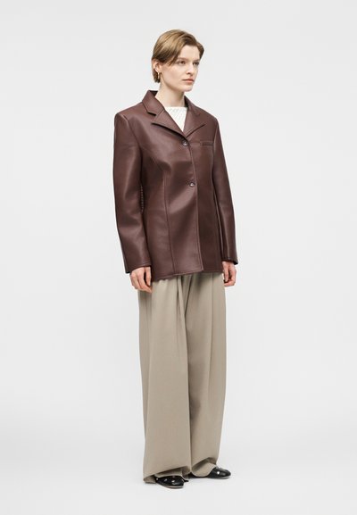 Άτομο που στέκεται φορώντας καφέ δερμάτινο στενό σακάκι, μπεζ παντελόνι φαρδιάς γραμμής, μαύρα φλατ παπούτσια και λευκό πλεκτό τοπ μπροστά σε απλό φόντο.