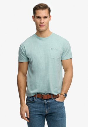 Joven con camiseta de manga corta y bolsillo de color azul claro, vaqueros azules, cinturón marrón y reloj de pulsera, de pie con una mano en el bolsillo.