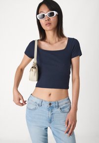 Námornícky pruhovaný crop top s krátkymi rukávmi, kombinovaný s svetlomodrými džínami. Modelka drží malú smotanovú kabelku a nosí biele obdĺžnikové slnečné okuliare.