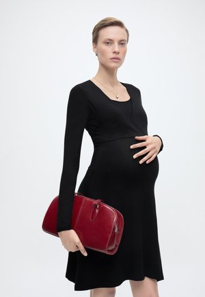 Nosečnica v črni obleki drži rdečo torbico in ima eno roko na trebuščku, postavljena na enobarvno svetlo ozadje.