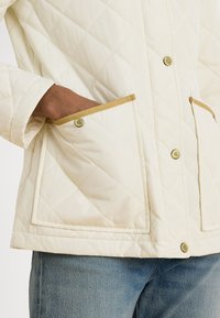 Geacă matlasată crem deschis, cu croială dreptunghiulară, cu garnitură aurie contrastantă la buzunare și nasturi, și o textură moale a materialului.