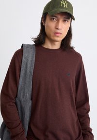 Brauner Strickpullover mit rundem Halsausschnitt und langen Ärmeln, kombiniert mit einer grauen Stoffschultertasche. Trägt eine grüne Cordmütze.