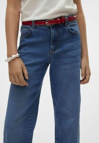 Jeans de mezclilla con un diseño de corte recto, lavado medio y costuras visibles, combinados con un cinturón de cuero sintético rojo brillante y una pulsera de aspecto perlado.