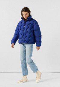 Blaues Pufferjackett mit diamantge quiltetem Muster, hohem Kragen und Reißverschluss; kombiniert mit hellblauen Jeans und beigen High-Top-Sneakern.