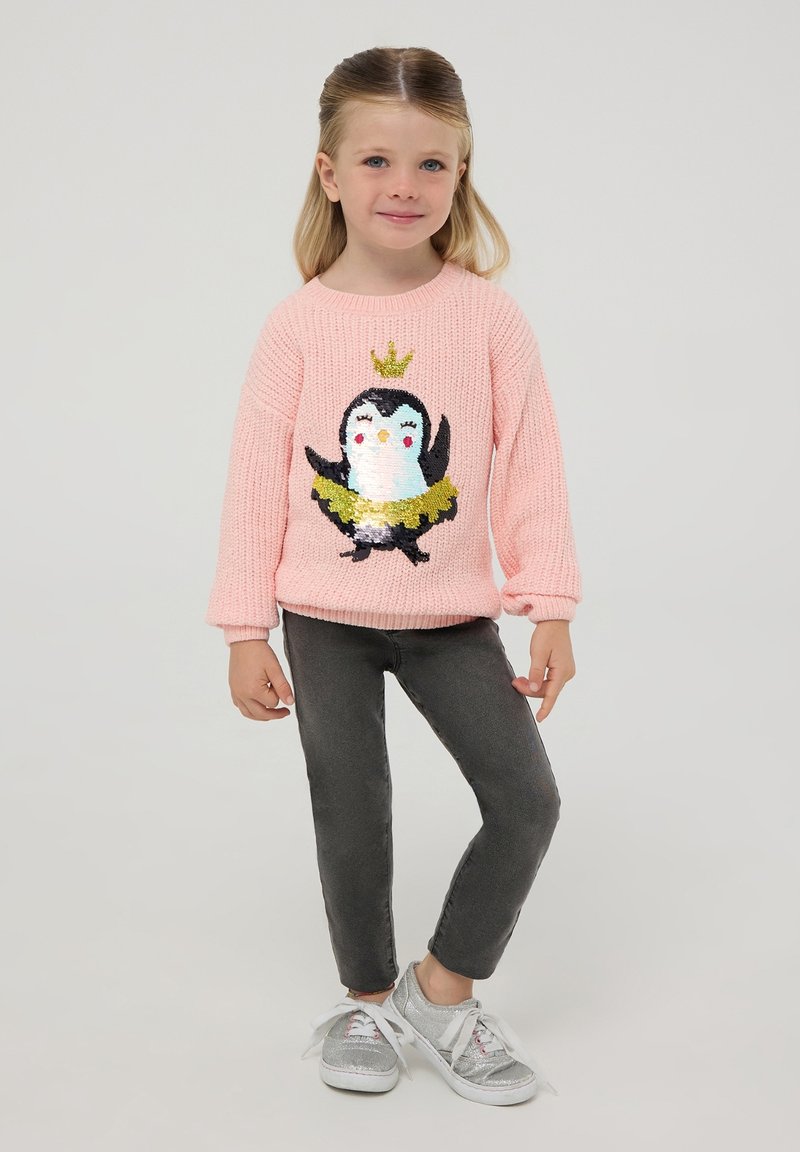 Maglione rosa lavorato a maglia con design di pinguino decorato con paillettes, abbinato a pantaloni aderenti grigi e scarpe argentate.