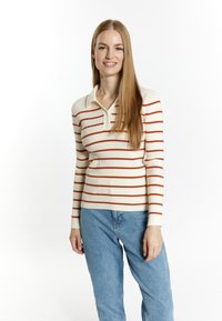 Junge Frau mit langen, glatten Haaren, die einen cremefarbenen Pullover mit orangefarbenen Streifen und blaue Jeans trägt, vor einem weißen Hintergrund stehend.