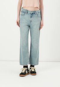 Jean large bleu clair à taille haute, avec fermeture à boutons, cinq poches et un aspect légèrement délavé, associé à des baskets noires.