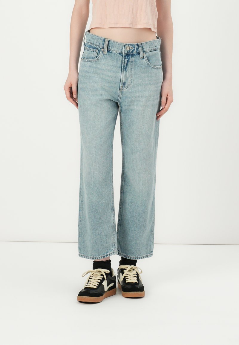 Jean large bleu clair à taille haute, avec fermeture à boutons, cinq poches et un aspect légèrement délavé, associé à des baskets noires.