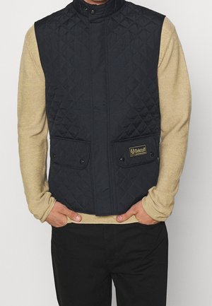 Man wearing a black quilted sleeveless vest over a beige long-sleeve shirt with hands tucked in front pockets.