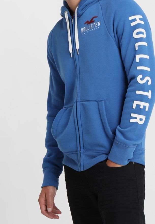 Blå zip-hoodie i mjukt tyg med vita dragsnören. Har "HOLLISTER" i fet vit text på ärmen och en liten logotyp på bröstet.