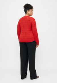 Rød strikket genser med et strukturert diamantmønster, rund hals og lange ermer, paret med svarte vide bukser og svart fottøy.