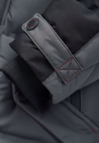 Gros plan sur le poignet d'une veste gris et noir avec une sangle ajustable et des coutures rouges visibles sur un tissu résistant.