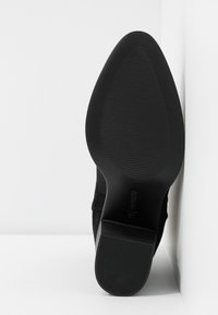 Semelle de chaussure noire à bout pointu avec motif antidérapant texturé, talon légèrement surélevé, positionnée contre un coin de mur et de sol blancs.