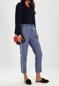Navy long-sleeve top, light blue trousers with white side stripe, and black slip-on shoes. Model holds a red and black clutch.