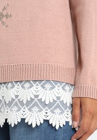 Pull en maille rose à ourlet côtelé sur un haut blanc orné de dentelle, associé à un jean bleu, main reposant près du côté.