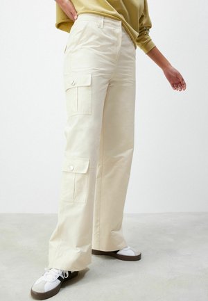 Personne portant un pantalon cargo beige à jambes larges avec poches à rabat et des baskets blanches avec semelles marron sur un fond clair.