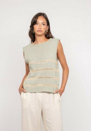 Femme portant un haut en maille sans manches vert clair avec des rayures horizontales beiges en ajouré et un pantalon beige ample, les mains dans les poches, fond neutre.