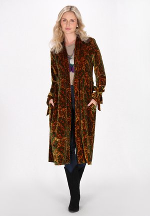 Lungo cappotto di velluto in un ricco motivo paisley arancione e verde, con apertura frontale, tasche laterali e maniche legate. Indossato sopra una maglietta casual e jeans.