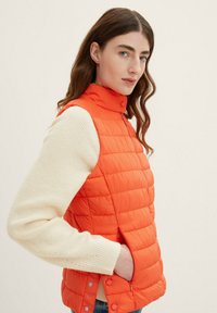 Helle orange Pufferweste mit hohem Kragen, Druckknöpfen und Seitentaschen. Getragen über einem cremefarbenen Strickpullover. Glatte Textur.