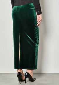 Gonna verde in velluto con un design a spacco sul retro, abbinata a scarpe nere con tacco alto. Il tessuto ha una consistenza morbida e lucida.