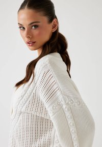 Hvid strikket cardigan med et struktureret åbent væv, intrikate kabelmønstre og v-halsdesign, med en subtil glans og blødt stof.