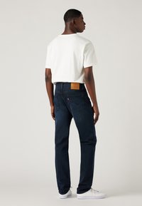 Egy férfi háttal áll, fehér pólót, sötétkék farmernadrágot bőrkéttel és fehér tornacipőt visel egy sima háttér előtt.