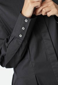 Fekete gombos ing, fitted mandzsettával, három ezüst gombbal, sima textúrával és pliszé részletekkel az ujjakon.