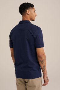 Námořnické modré polo tričko s krátkým rukávem a límečkem, hladká textura a detail švu na zadním rameni. Nosené s béžovými kalhotami.
