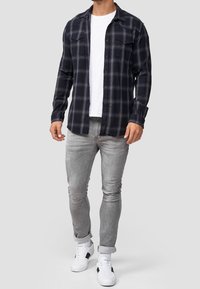 Mies, który ma na sobie czarną i szaro-skalną koszulę w kratę na białym t-shircie, jasnoszare dżinsy i białe sneakersy z czarnymi akcentami na jednolitym tle.