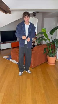 Sproščena obleka z temno modrim zip-up jopičem, belim majico, ohlapnimi črnimi hlačami in srebrnimi športnimi copati na lesenem tleh, obdanimi s rastlinami.