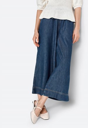 Wide-leg denim culottes in dark blue with stitched hems, paired with light beige slingback flats. Soft texture and relaxed fit.