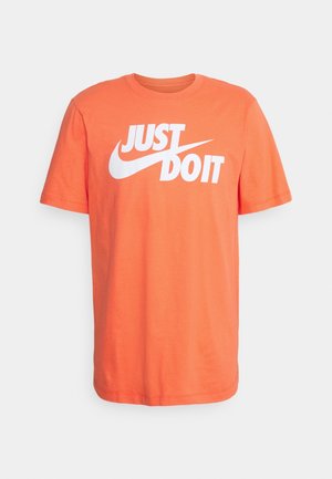 Pomarańczowy T-shirt z krótkim rękawem z białym napisem "JUST DO IT" i logo Nike Swoosh na klatce piersiowej na jednolitym tle.