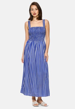 Woman standing wearing a blue and white vertical striped sleeveless maxi dress with smocked bodice and white open-toe sandals.