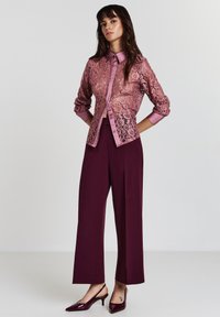 Femme se tient avec les mains derrière le dos, portant un blouse en dentelle rose transparent, un pantalon bordeaux à taille haute et jambes larges, ainsi que des talons aiguilles bordeaux à brides.