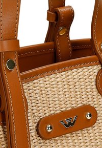 Splotkowana torba z trawy ze skórzanymi uchwytami i akcentami w kolorze tan. Posiada złotą tabliczkę z logo oraz detale wyszywane. Kompaktowy i usystematyzowany design.