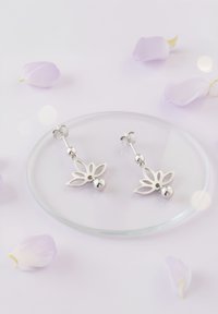 Silverörhängen med en blomsterutskärning och runda pärl-accenter, placerade på en glasfat omgiven av bleka blomblad.